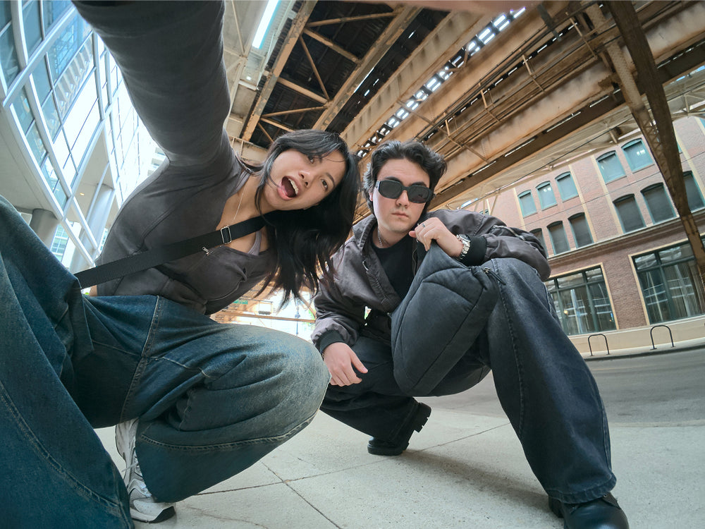 Two friends laughing under an elevated train, one hanging off a railing, both wearing Bottle Bag crossbody