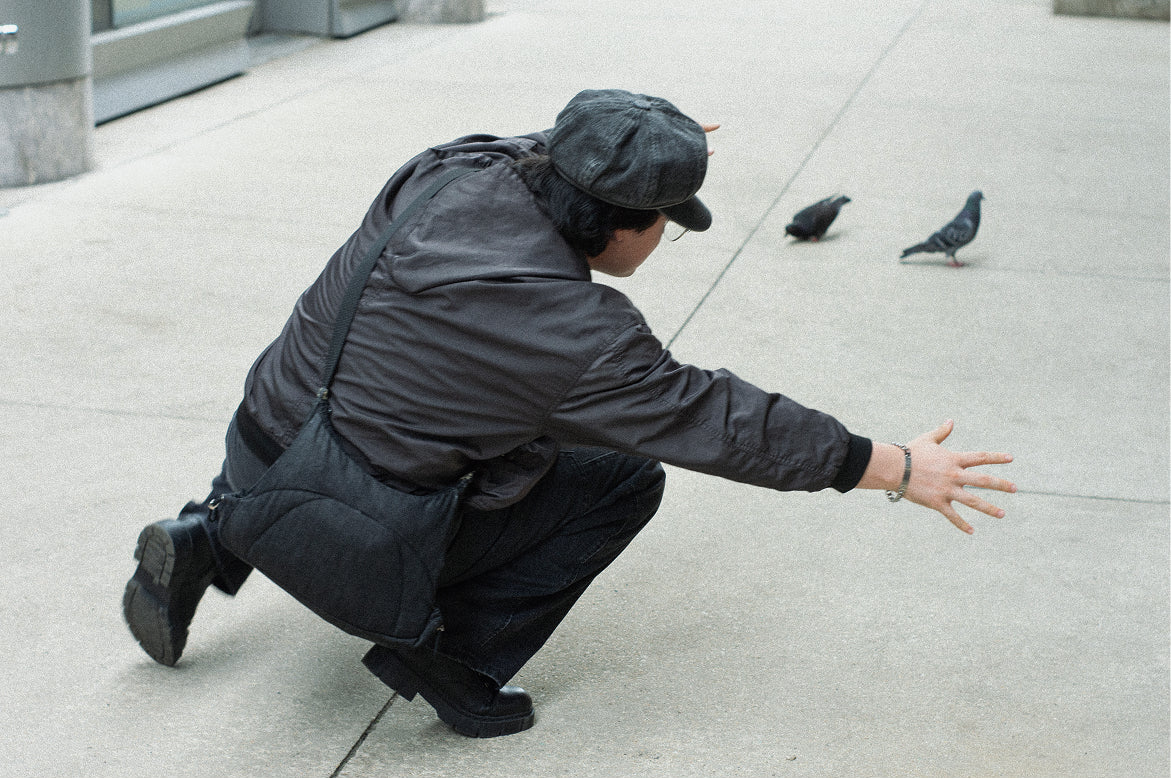Person in a dark jacket and cap crouching low on a sidewalk toward two pigeons, Bottle Bag worn crossbody
