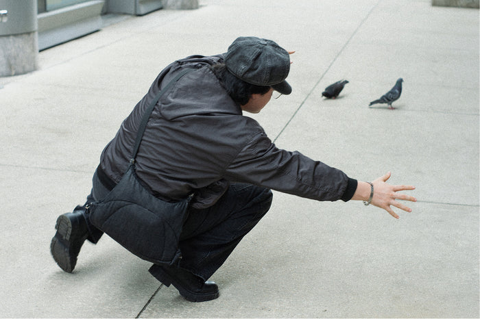 Person in a dark jacket and cap crouching low on a sidewalk toward two pigeons, Bottle Bag worn crossbody