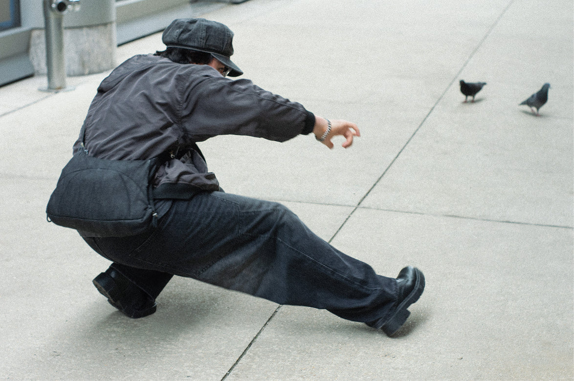 Person in a dark jacket and cap crouched on a sidewalk reaching toward pigeons, Bottle Bag on their back
